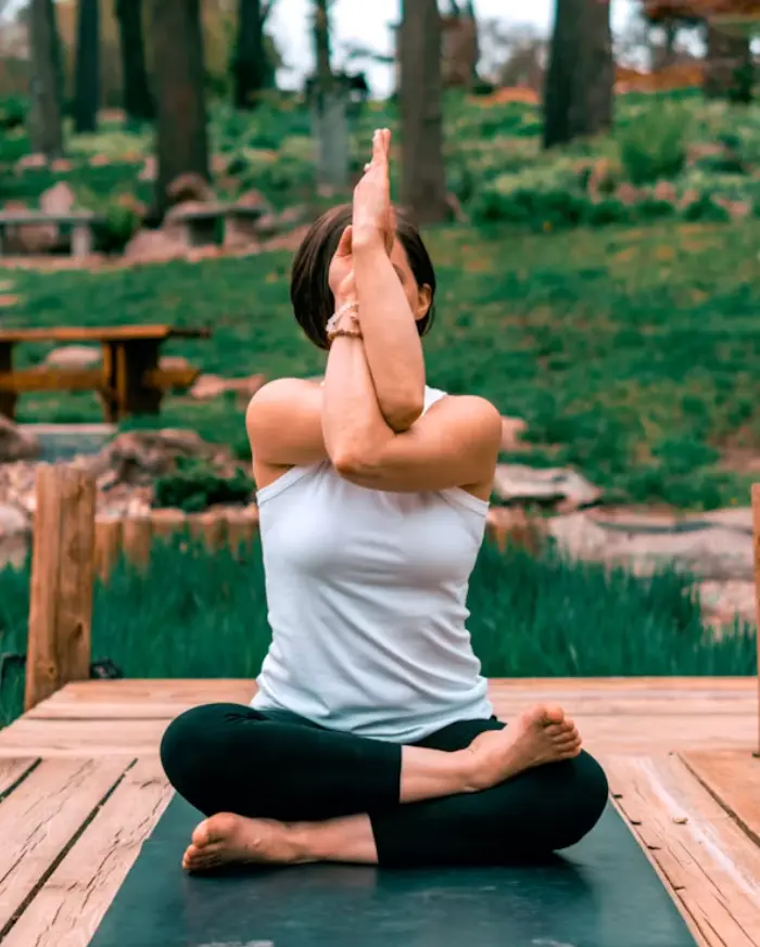 Yoga practitioner in eagle arms pose on a wooden deck surrounded by greenery